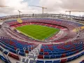 Widok na budowane nowe Camp Nou z zieloną murawą i czerwono-niebieskimi trybunami. W tle widać dźwigi budowlane.