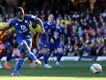 Piłkarz Leicester w niebieskiej koszulce strzela do bramki w meczu Leicester – Watford. W tle kibice i zawodnicy Watford w żółto-czerwonych strojach.