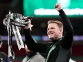 Trener z radością unosi puchar ligi angielskiej. Zawodnicy świętują zwycięstwo, a trofeum lśni w blasku świateł.