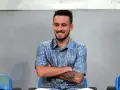 Uśmiechnięty Afonso Sousa w niebieskiej koszuli z wzorem Diora, siedzi ze skrzyżowanymi ramionami.