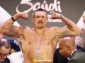 Ołeksandr Usyk, bokser, napina bicepsy, prezentując tatuaże i krzyż na łańcuchu. W tle widać innych mężczyzn i pas mistrzowski.