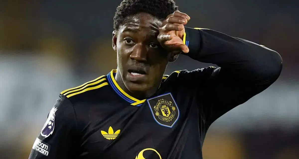 Młody piłkarz Kobbie Mainoo w czarnej koszulce Manchester United z żółtymi pasami, wyciera pot z czoła.