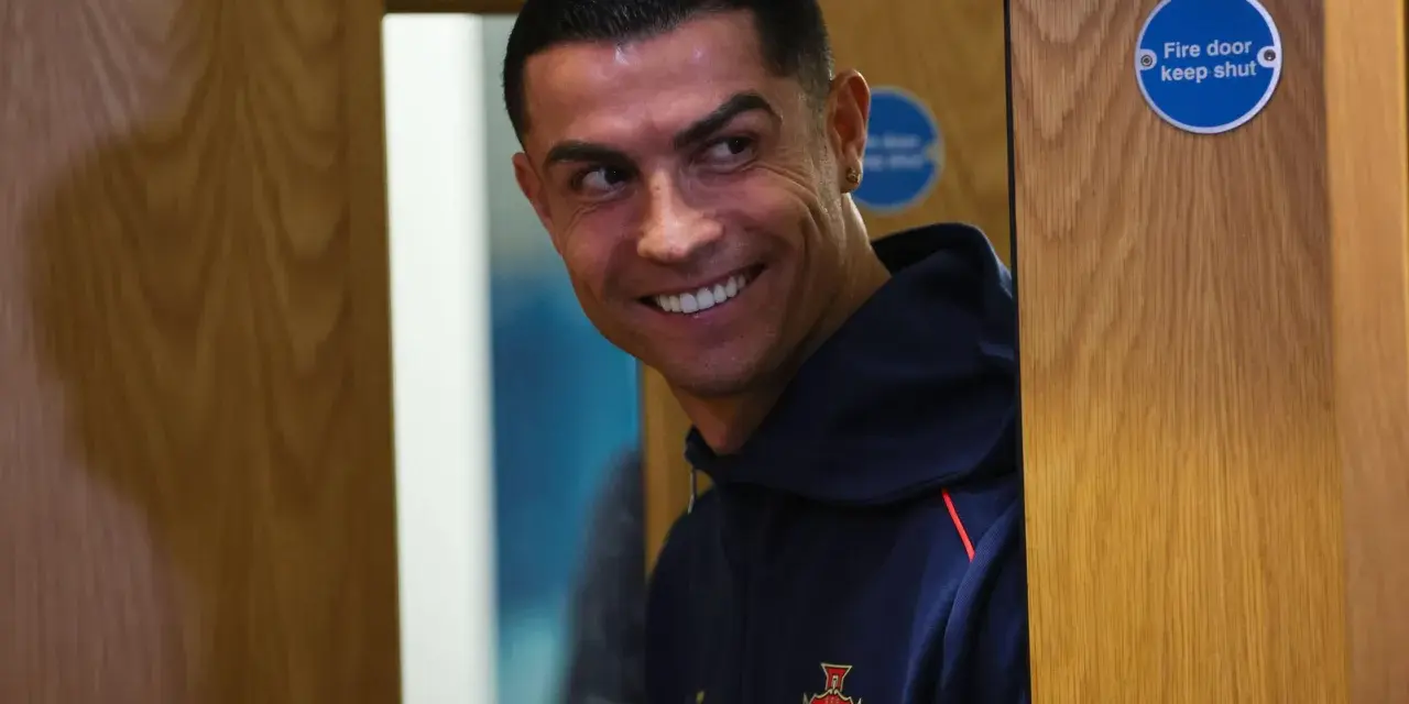 Uśmiechnięty Cristiano Ronaldo w granatowej bluzie z herbem Portugalii. Zastanawia się nad datami dołączenia do drużyny.