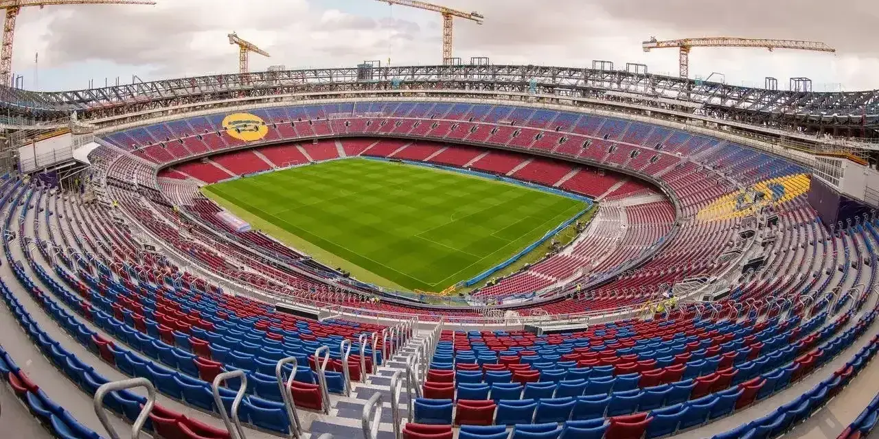 Widok na budowane nowe Camp Nou z zieloną murawą i czerwono-niebieskimi trybunami. W tle widać dźwigi budowlane.