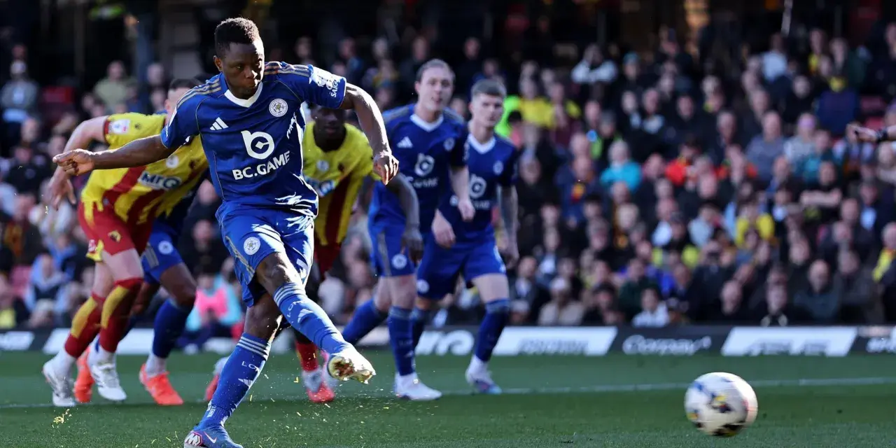 Piłkarz Leicester w niebieskiej koszulce strzela do bramki w meczu Leicester – Watford. W tle kibice i zawodnicy Watford w żółto-czerwonych strojach.