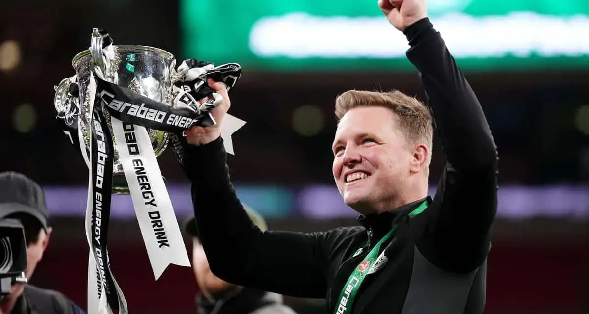 Trener z radością unosi puchar ligi angielskiej. Zawodnicy świętują zwycięstwo, a trofeum lśni w blasku świateł.
