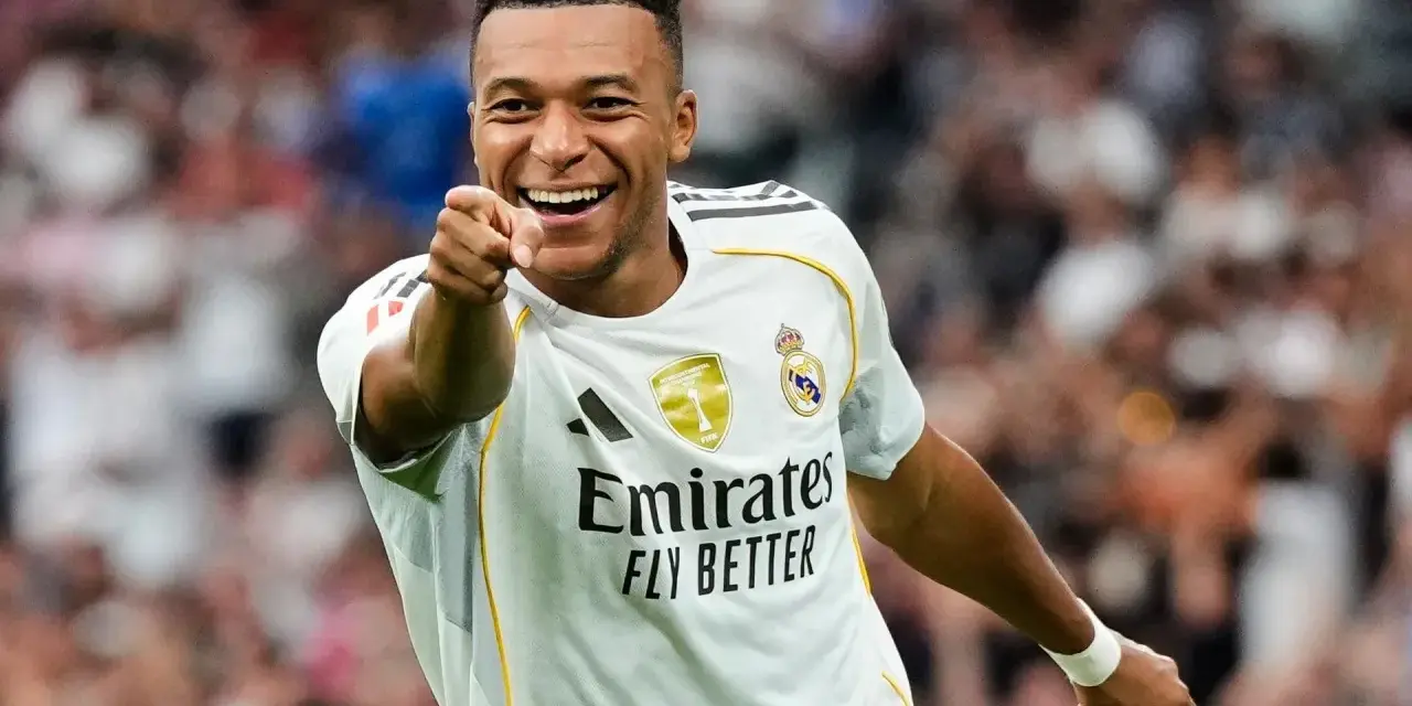 Uśmiechnięty Kylian Mbappé w białej koszulce Realu Madryt wskazuje palcem na widza, celebrując zwycięstwo.