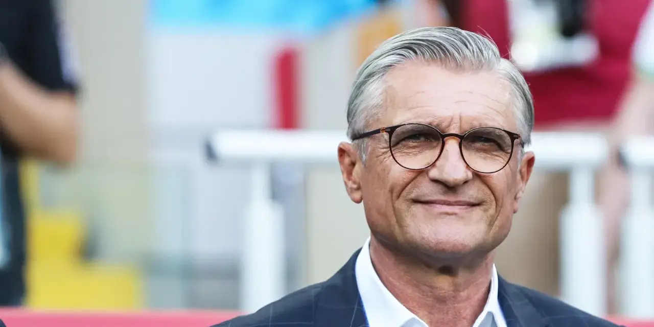 Uśmiechnięty mężczyzna w okularach, były trener reprezentacji Polski, w garniturze i białej koszuli.