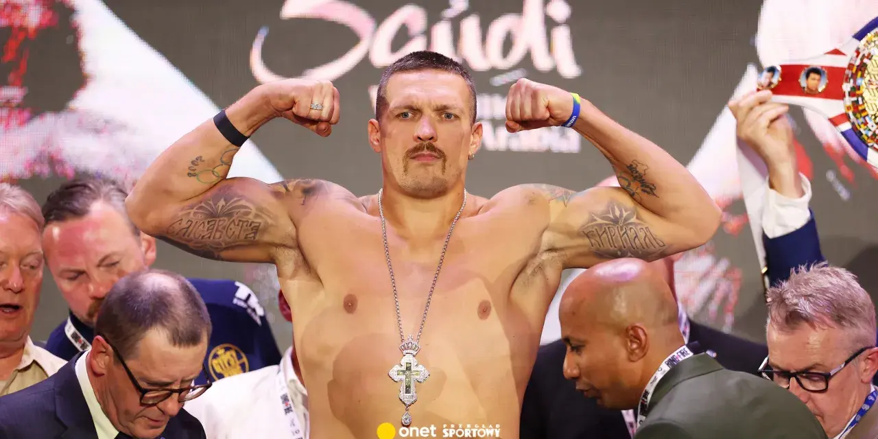 Ołeksandr Usyk, bokser, napina bicepsy, prezentując tatuaże i krzyż na łańcuchu. W tle widać innych mężczyzn i pas mistrzowski.