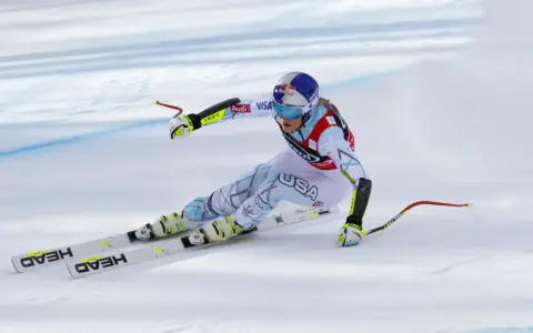 Lindsey Vonn w pełnym pędzie, w białym kombinezonie z napisem USA, na nartach HEAD. Widoczny moment tuż przed upadkiem.