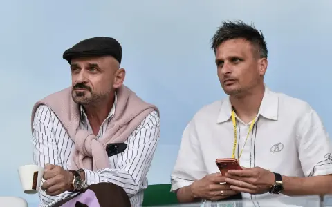 Dwóch mężczyzn siedzi na trybunach. Jeden w czapce i swetrze na ramionach, drugi z telefonem. Na smyczy widnieje napis KS Wieczysta Kraków skład.