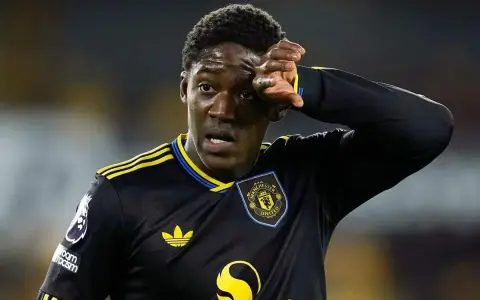 Młody piłkarz Kobbie Mainoo w czarnej koszulce Manchester United z żółtymi pasami, wyciera pot z czoła.