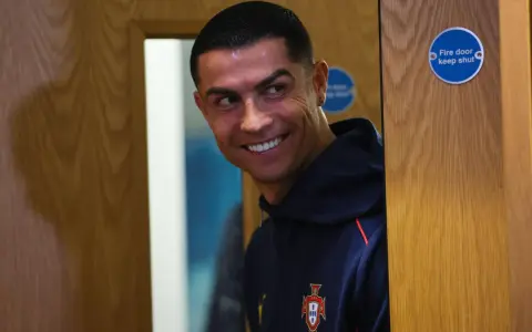 Uśmiechnięty Cristiano Ronaldo w granatowej bluzie z herbem Portugalii. Zastanawia się nad datami dołączenia do drużyny.