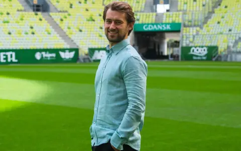 Uśmiechnięty Tomasz Kaczmarek, trener, stoi na tle stadionu w Gdańsku.