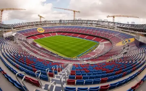 Widok na budowane nowe Camp Nou z zieloną murawą i czerwono-niebieskimi trybunami. W tle widać dźwigi budowlane.