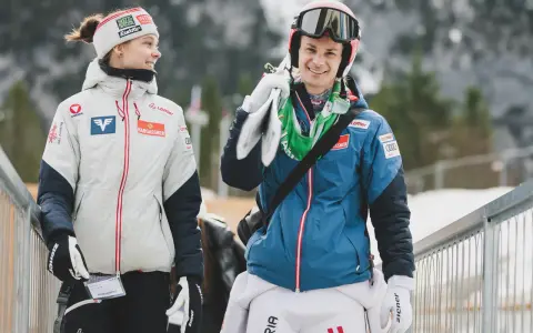 Dwóch zawodników mś skoki narciarskie, młoda kobieta w białej kurtce i mężczyzna w niebieskiej, niosący narty.