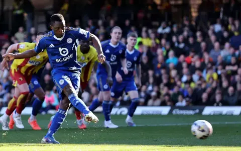 Piłkarz Leicester w niebieskiej koszulce strzela do bramki w meczu Leicester – Watford. W tle kibice i zawodnicy Watford w żółto-czerwonych strojach.