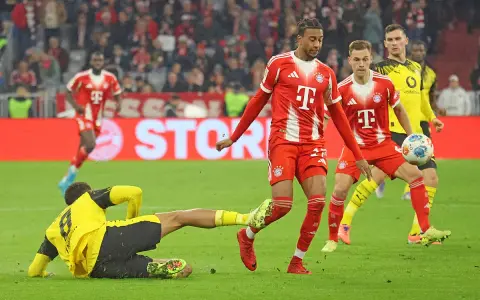 Intensywna akcja na boisku podczas meczu składy: Borussia Dortmund – Bayern Monachium. Piłkarz Bayernu w czerwonej koszulce próbuje odebrać piłkę leżącemu zawodnikowi BVB.