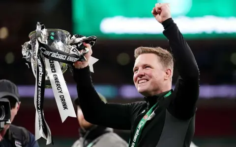 Trener z radością unosi puchar ligi angielskiej. Zawodnicy świętują zwycięstwo, a trofeum lśni w blasku świateł.