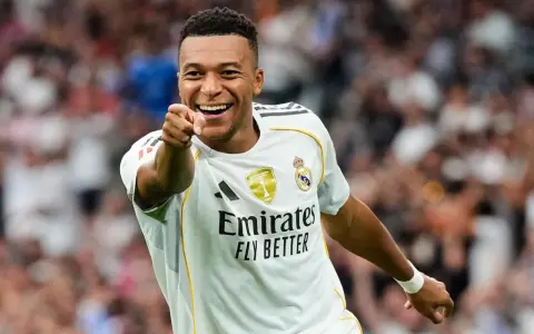 Uśmiechnięty Kylian Mbappé w białej koszulce Realu Madryt wskazuje palcem na widza, celebrując zwycięstwo.