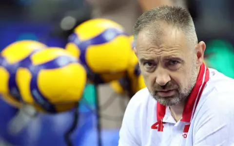 Skupiony Nikola Grbić, trener siatkówki, w białej koszulce z czerwonymi akcentami, na tle żółto-niebieskich piłek.