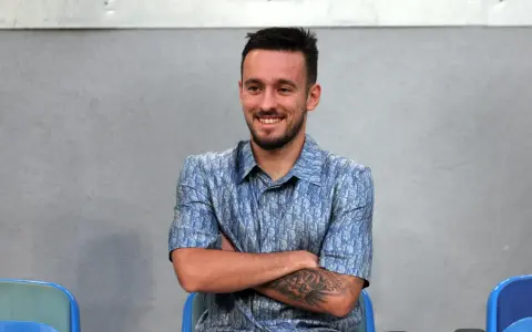 Uśmiechnięty Afonso Sousa w niebieskiej koszuli z wzorem Diora, siedzi ze skrzyżowanymi ramionami.