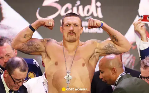Ołeksandr Usyk, bokser, napina bicepsy, prezentując tatuaże i krzyż na łańcuchu. W tle widać innych mężczyzn i pas mistrzowski.