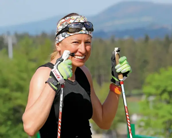 Uśmiechnięta Justyna Kowalczyk-Tekieli z kijkami do nartorolkarstwa, w sportowym stroju i opasce na głowie, gotowa do treningu.