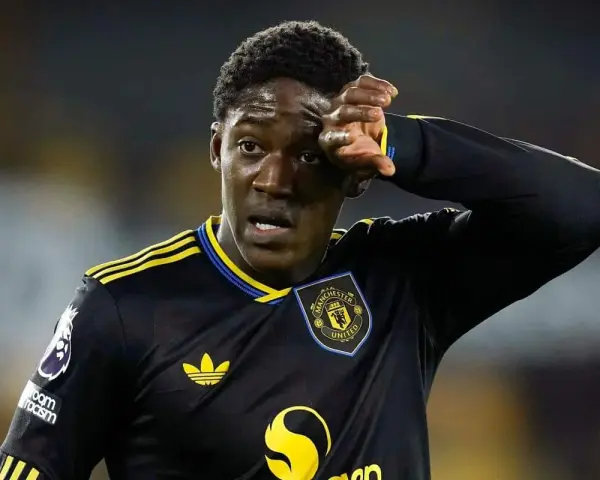 Młody piłkarz Kobbie Mainoo w czarnej koszulce Manchester United z żółtymi pasami, wyciera pot z czoła.