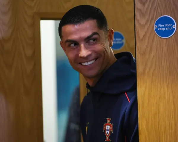 Uśmiechnięty Cristiano Ronaldo w granatowej bluzie z herbem Portugalii. Zastanawia się nad datami dołączenia do drużyny.