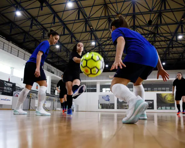 Graczki w hali sportowej ćwiczą zagrania, jedna z nich wykonuje pivot, podając piłkę nożną.
