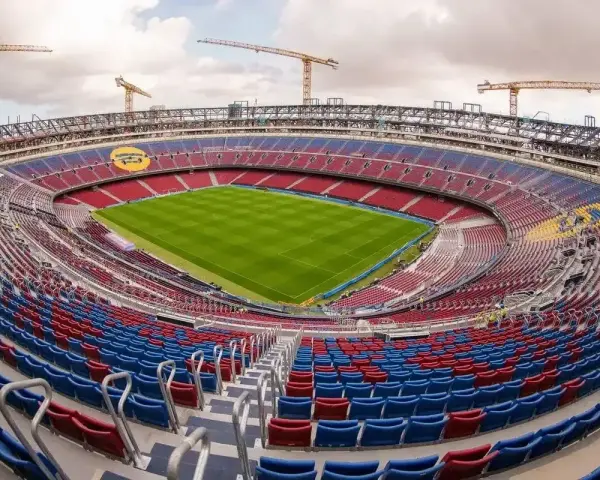 Widok na budowane nowe Camp Nou z zieloną murawą i czerwono-niebieskimi trybunami. W tle widać dźwigi budowlane.