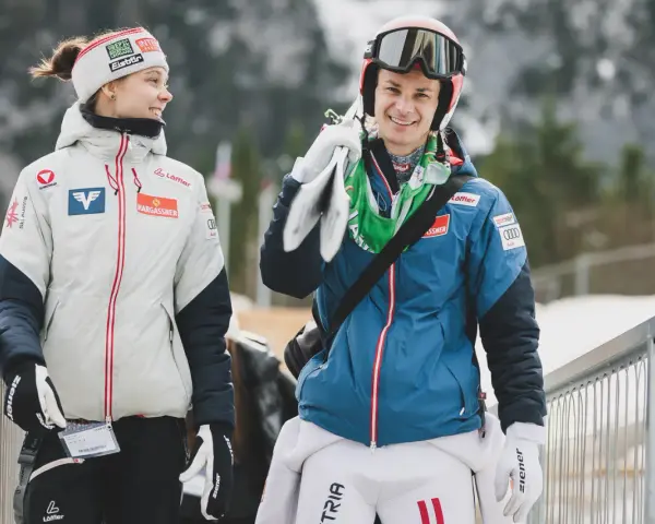 Dwóch zawodników mś skoki narciarskie, młoda kobieta w białej kurtce i mężczyzna w niebieskiej, niosący narty.
