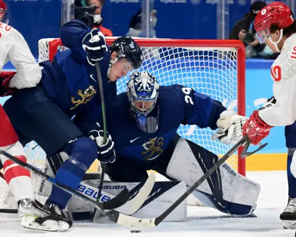 Bramkarz Finlandii broni strzał. W tle zawodnicy walczą o krążek, dążąc do zwycięstwa, jak przystało na mistrzów olimpijskich w hokeju na lodzie.