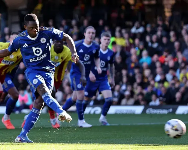 Piłkarz Leicester w niebieskiej koszulce strzela do bramki w meczu Leicester – Watford. W tle kibice i zawodnicy Watford w żółto-czerwonych strojach.