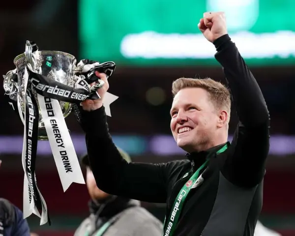 Trener z radością unosi puchar ligi angielskiej. Zawodnicy świętują zwycięstwo, a trofeum lśni w blasku świateł.