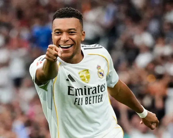 Uśmiechnięty Kylian Mbappé w białej koszulce Realu Madryt wskazuje palcem na widza, celebrując zwycięstwo.