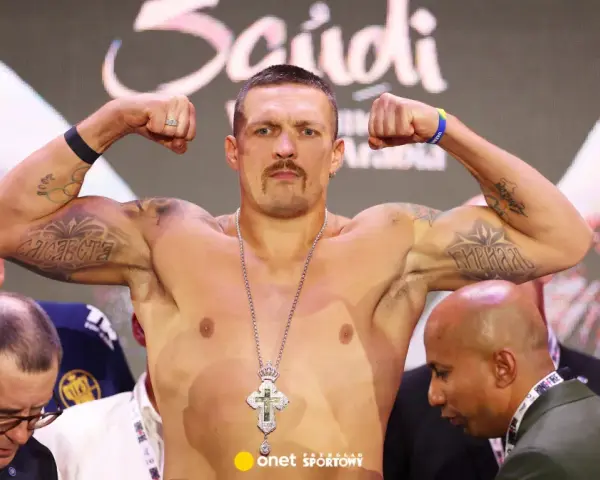 Ołeksandr Usyk, bokser, napina bicepsy, prezentując tatuaże i krzyż na łańcuchu. W tle widać innych mężczyzn i pas mistrzowski.