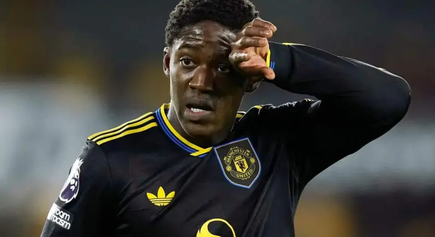 Młody piłkarz Kobbie Mainoo w czarnej koszulce Manchester United z żółtymi pasami, wyciera pot z czoła.