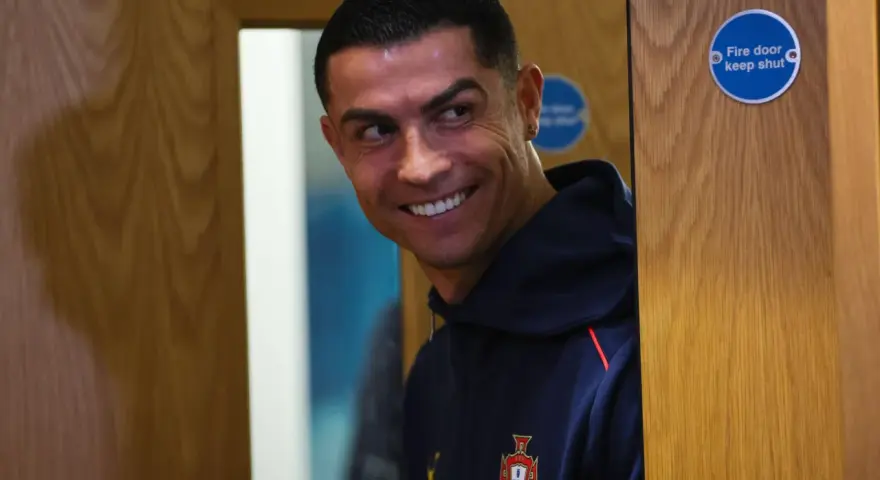 Uśmiechnięty Cristiano Ronaldo w granatowej bluzie z herbem Portugalii. Zastanawia się nad datami dołączenia do drużyny.