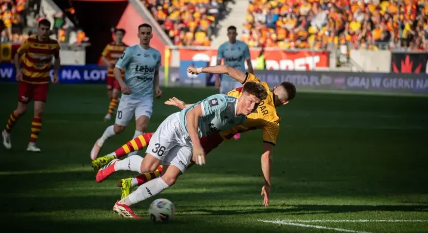 Zacięta walka na boisku podczas meczu Jagiellonia Białystok – Piast. Piłkarze walczą o piłkę, jeden z nich upada.