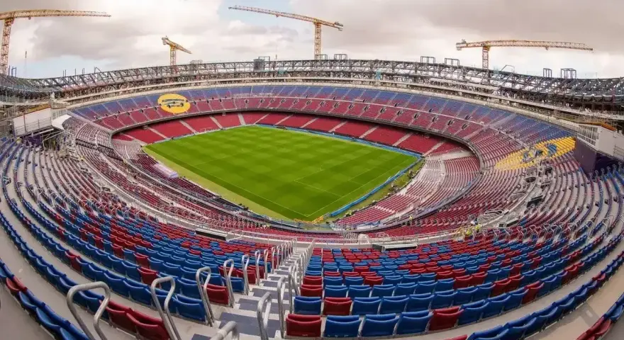 Widok na budowane nowe Camp Nou z zieloną murawą i czerwono-niebieskimi trybunami. W tle widać dźwigi budowlane.