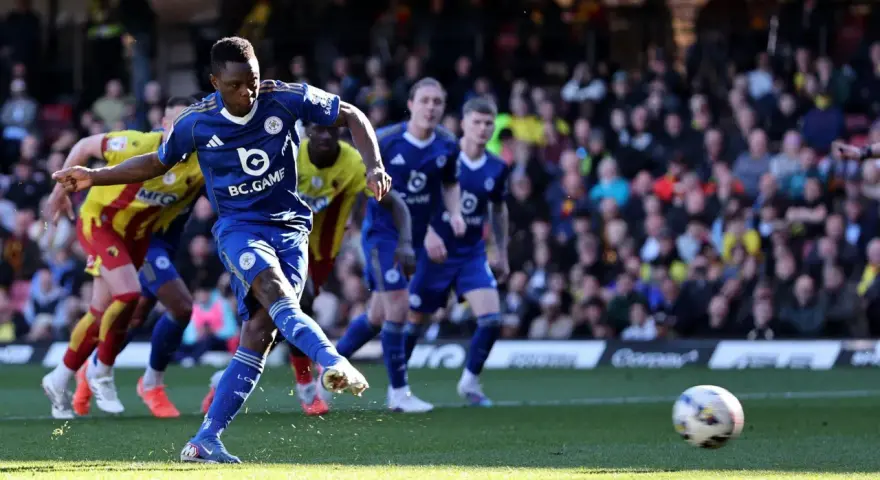 Piłkarz Leicester w niebieskiej koszulce strzela do bramki w meczu Leicester – Watford. W tle kibice i zawodnicy Watford w żółto-czerwonych strojach.