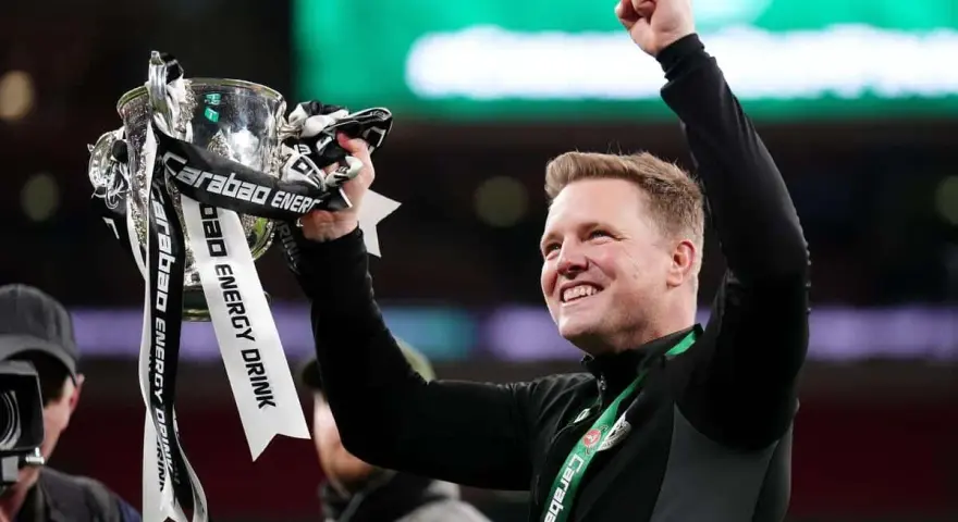 Trener z radością unosi puchar ligi angielskiej. Zawodnicy świętują zwycięstwo, a trofeum lśni w blasku świateł.