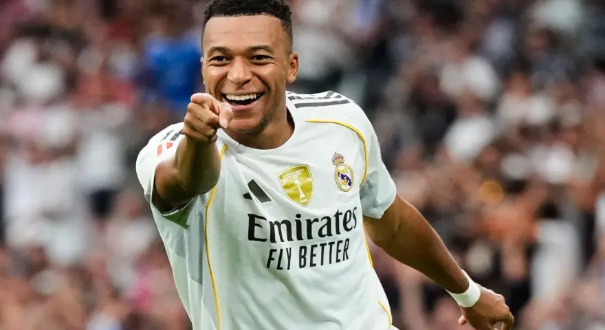 Uśmiechnięty Kylian Mbappé w białej koszulce Realu Madryt wskazuje palcem na widza, celebrując zwycięstwo.