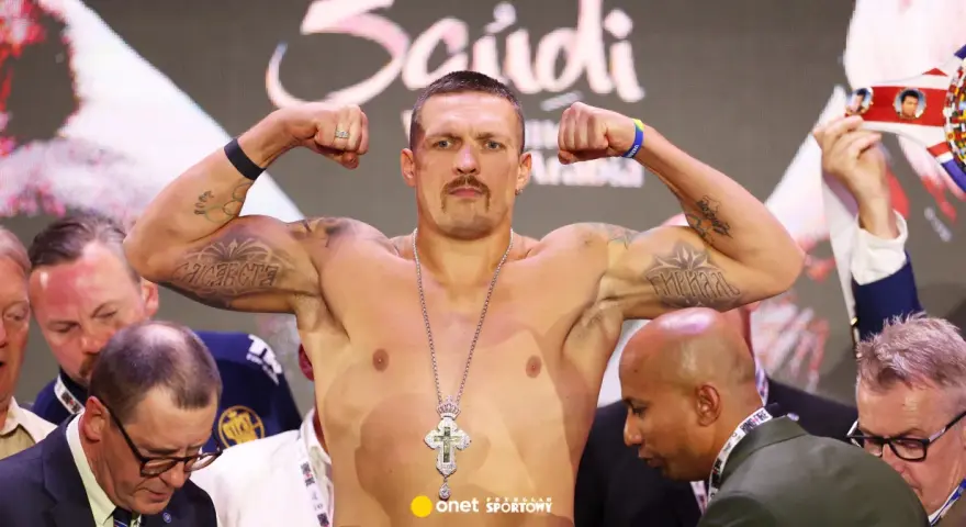 Ołeksandr Usyk, bokser, napina bicepsy, prezentując tatuaże i krzyż na łańcuchu. W tle widać innych mężczyzn i pas mistrzowski.