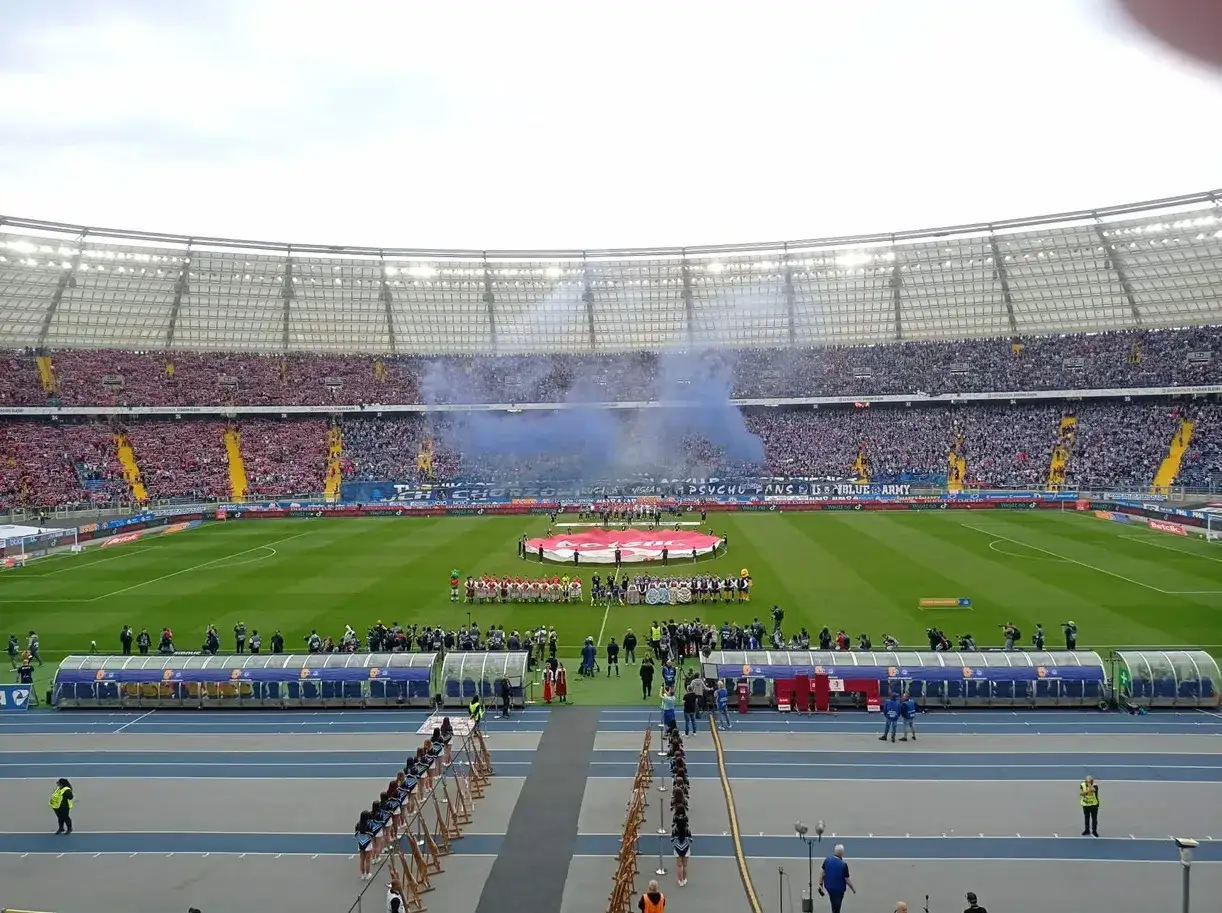 Piłkarze na murawie stadionu, dymią race, trybuny pełne kibiców. Zapowiada się emocjonujący mecz, a wyniki i ligi piłki nożnej będą śledzone z zapartym tchem.