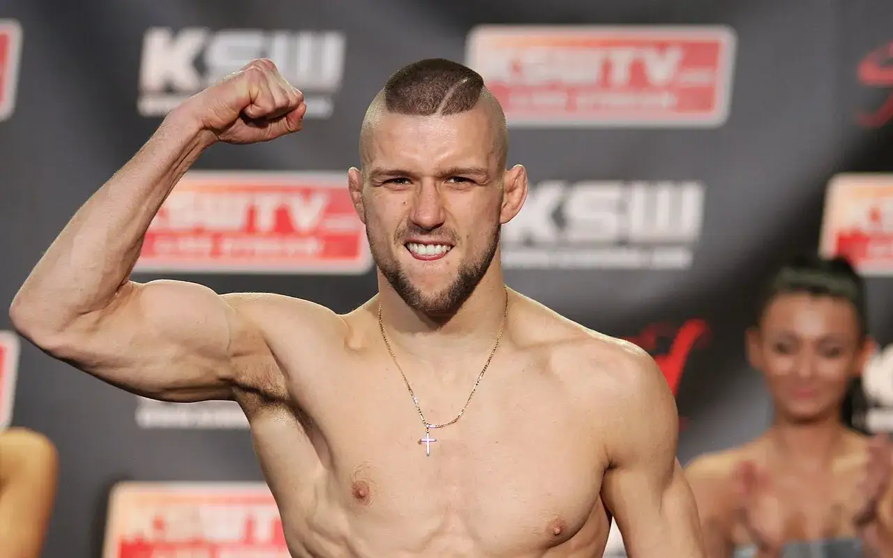 Młody zawodnik MMA z charakterystyczną fryzurą i zarostem napina biceps. W tle logo KSW, symbolizuje moment, kiedy walczy gamrot.