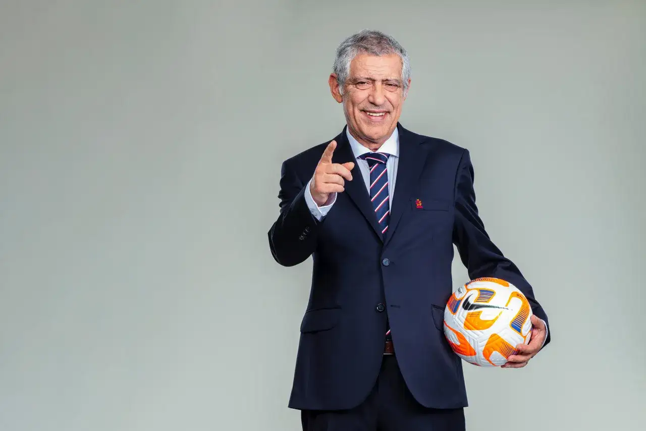 Uśmiechnięty Fernando Santos w garniturze wskazuje palcem i trzyma piłkę nożną.