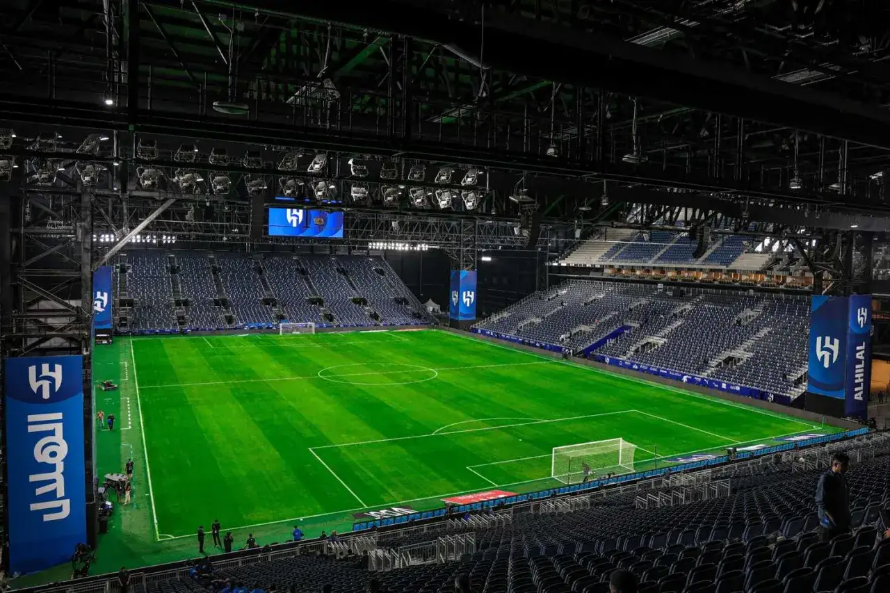 Pusta, zielona murawa stadionu piłkarskiego Al Hilal, z rzędami niebieskich siedzeń i oświetleniem.